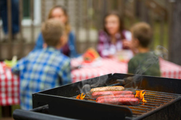 a hot grill with several hot dogs in the foreground, with a family of four seated around a picnic table in the background out of focus